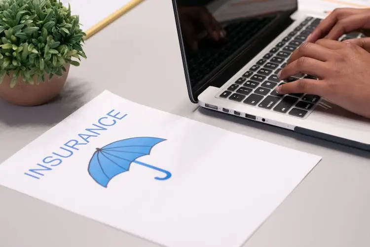 A person typing on a white laptop, paper with an umbrella and the word Insurance next to it.